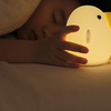 Twinkle Glory silicone cuckoo night light glowing with warm yellow light next to a sleeping child in bed