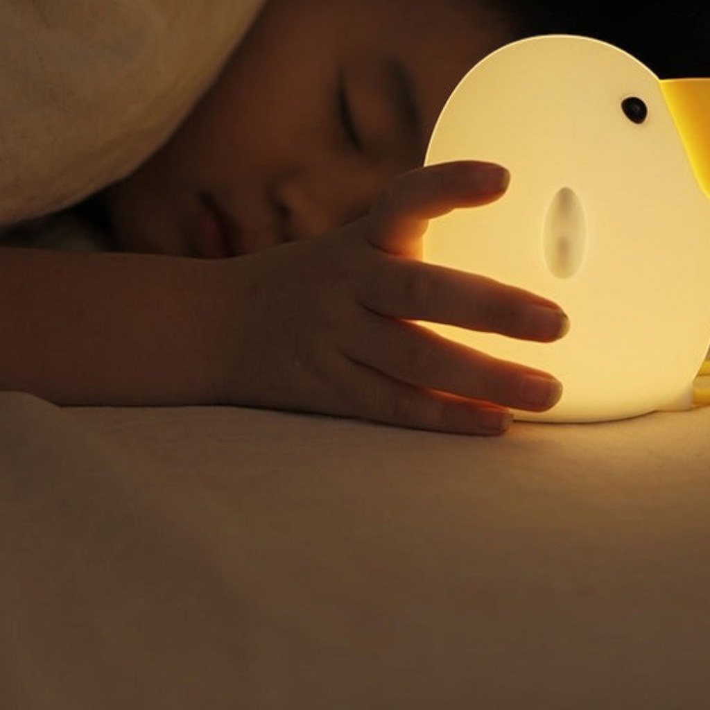 Twinkle Glory silicone cuckoo night light glowing with warm yellow light next to a sleeping child in bed