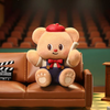Plush toy bear in a formal outfit sitting on a brown leather couch with a clapperboard in an auditorium setting.