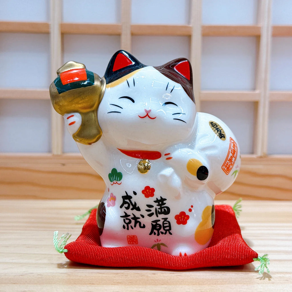 A white ceramic Japanese maneki-neko lucky cat figurine with a raised gold paw, sitting on a red fabric cushion. The cat has black and red markings, with Japanese characters painted on its belly, set against a light wooden background.