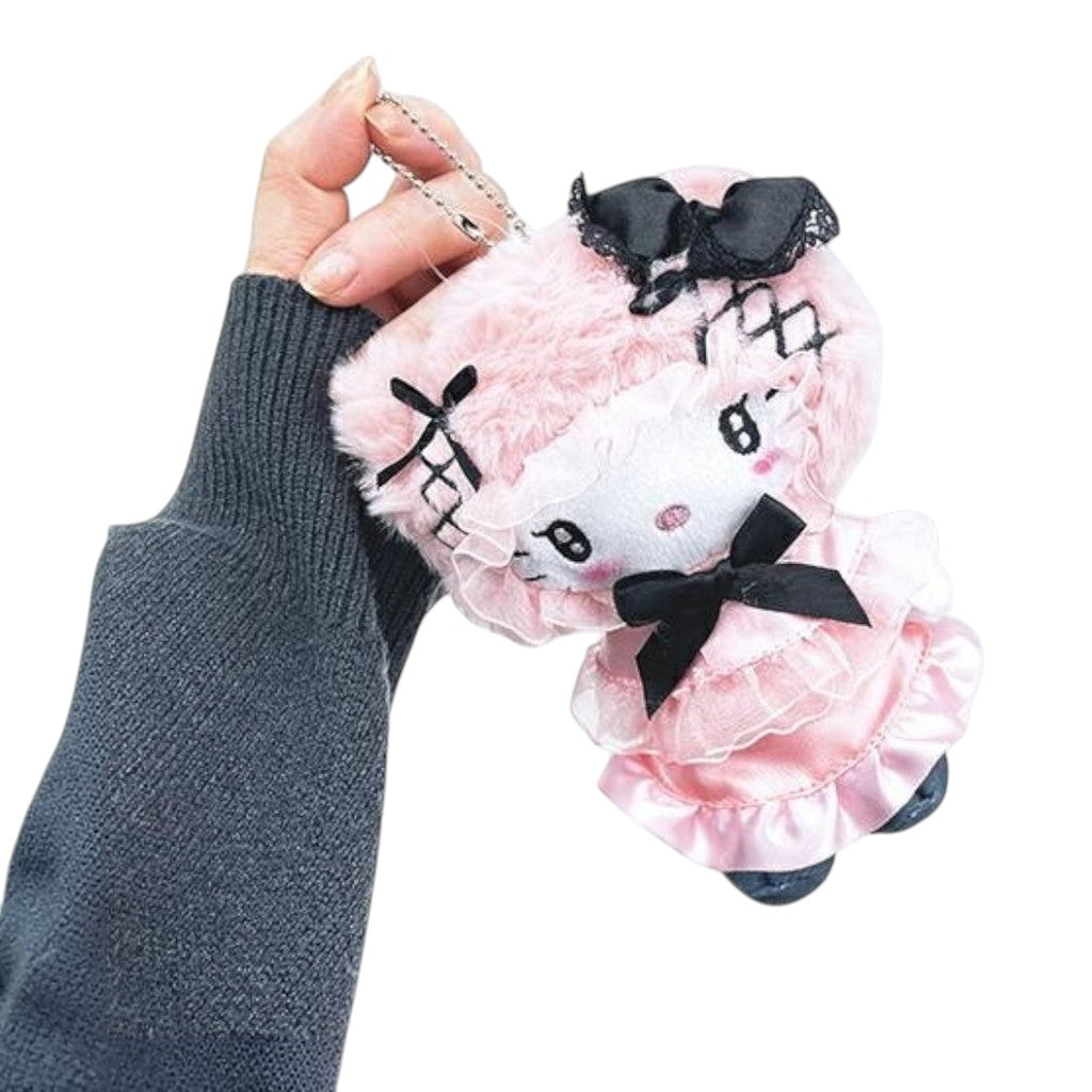 A hand wearing a dark gray knit sweater holds a pink plush doll keychain with black bows and lace details against a plain white background.