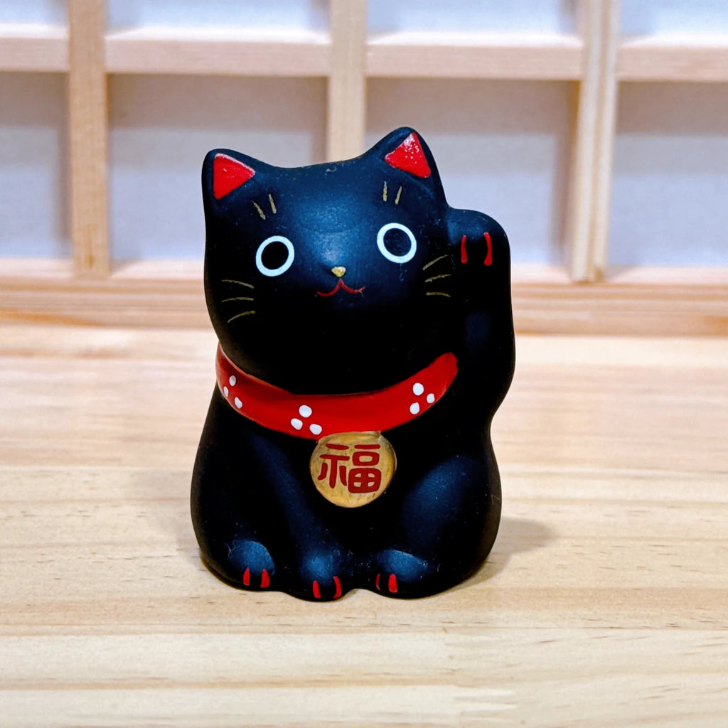 A black maneki-neko lucky cat figurine with a raised paw, wearing a red collar with white dots and a gold medallion, set on a light wood surface.
