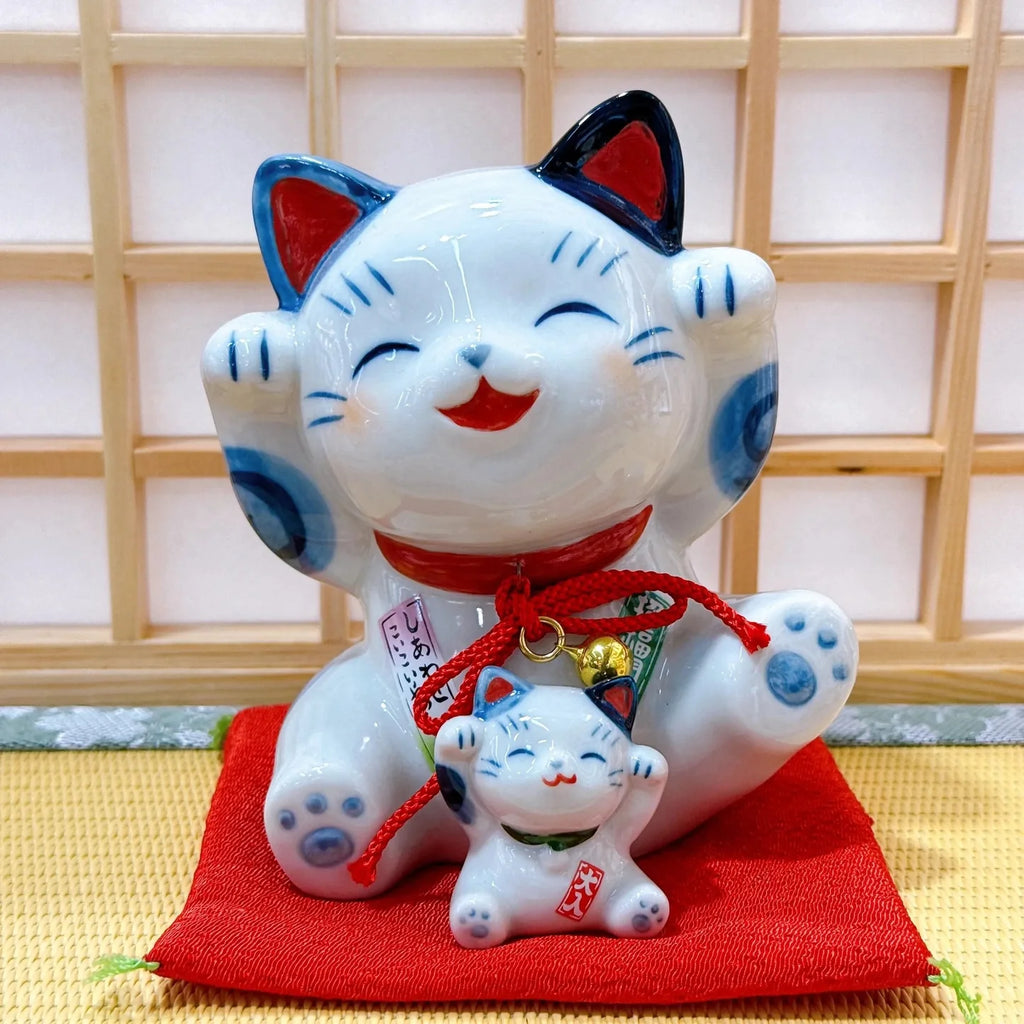 White cat figurine with blue accents and red string on a red cushion against a wooden panel background.