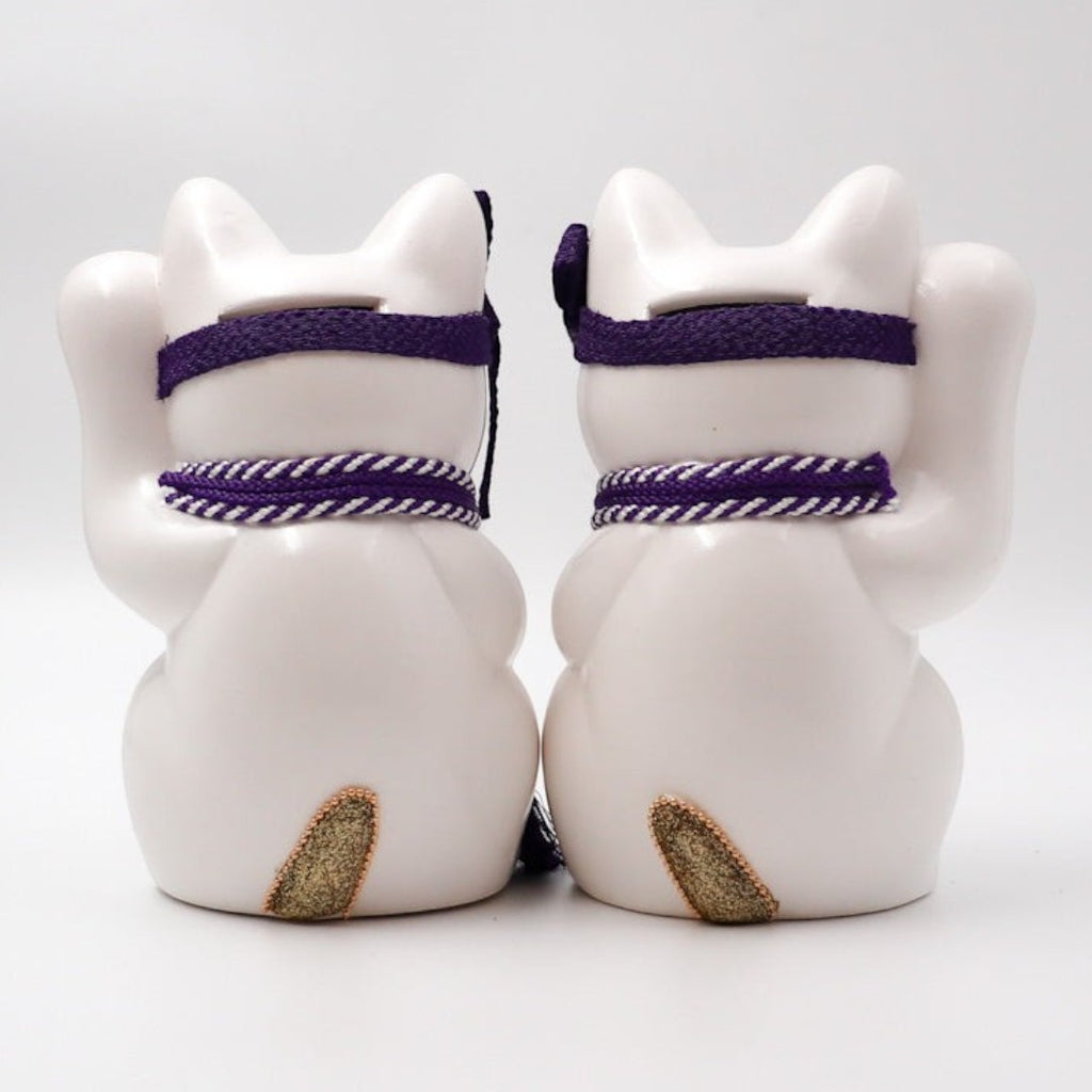 Back view of two Twinkle Glory Goenneko ceramic lucky cats with purple headbands and gold tails on a white background