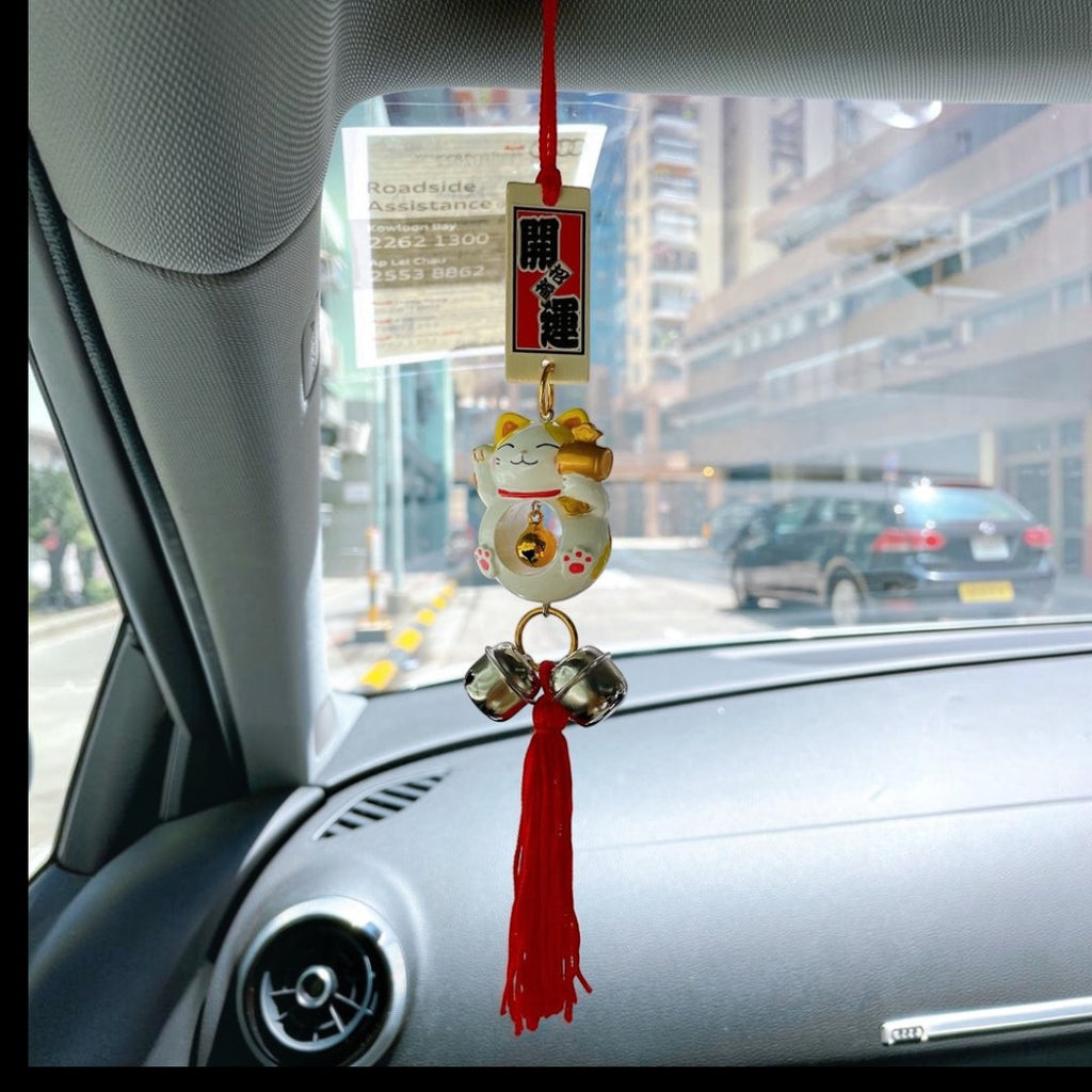 Twinkle Glory Japanese lucky cat car charm with a waving paw, gold bell, and red tassel hanging from a rearview mirror