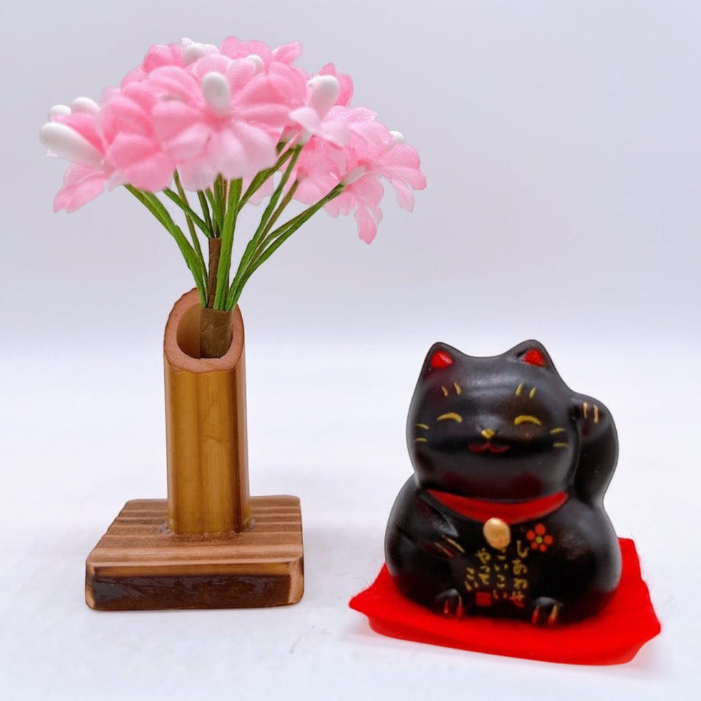 Twinkle Glory Yakushigama mini black lucky cat figurine on a red mat with a small pink flower arrangement