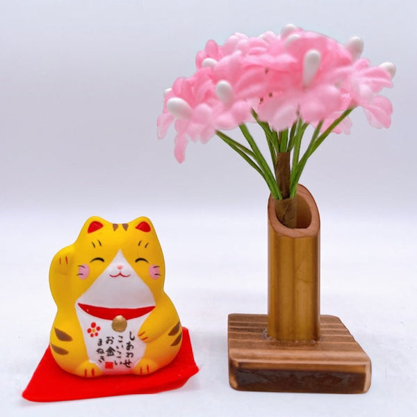 Twinkle Glory Yakushigama mini yellow lucky cat figurine on a red mat next to a small bamboo vase with pink flowers