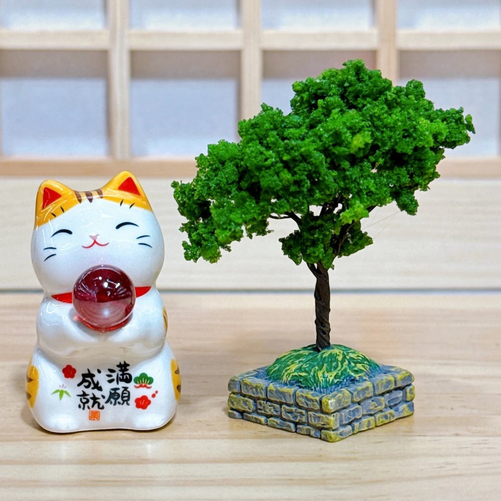 Twinkle Glory Yakushigama porcelain lucky cat figurine holding a red orb next to a miniature green tree model